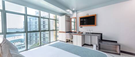 Habitación de hotel moderna con una cama grande, un escritorio, un televisor y vistas de la ciudad a través de amplias ventanas.