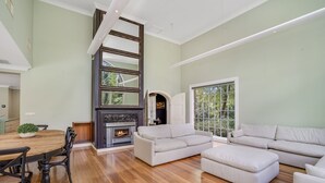A spacious living room with a fireplace, a dining area with a wooden table and chairs, and a large window providing natural light. - Karinya Parkland Estate (New Lambton Heights)
