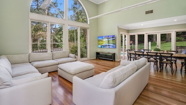 A spacious living room with a large sectional sofa, a dining area with a wooden table and chairs, and a TV mounted on the wall. - Karinya Parkland Estate (New Lambton Heights)