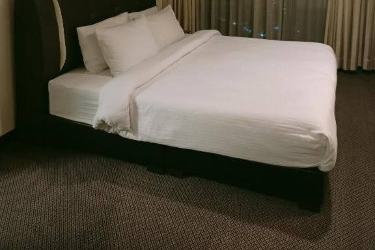 A hotel room with a large bed, a headboard, a window with curtains, a wall-mounted air vent, and a framed picture on the wall.