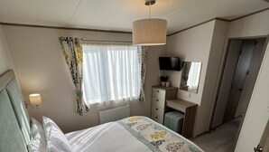 Ein Schlafzimmer mit Bett, Nachttischlampe, Fenster mit Vorhängen, Schreibtisch mit Stuhl und einem Fernseher.