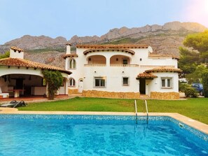 Eine große, weiße Villa mit Swimmingpool, umgeben von einem gepflegten Rasen und einem Bergpanorama.