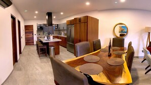 A modern kitchen with a central island, wooden cabinets, and a dining area with a large wooden table and chairs. - Bahía #304, Sabina Residences (Cabo San Lucas)