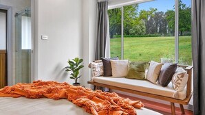 A bedroom with a large window, a bed, a bench with cushions, and a potted plant.