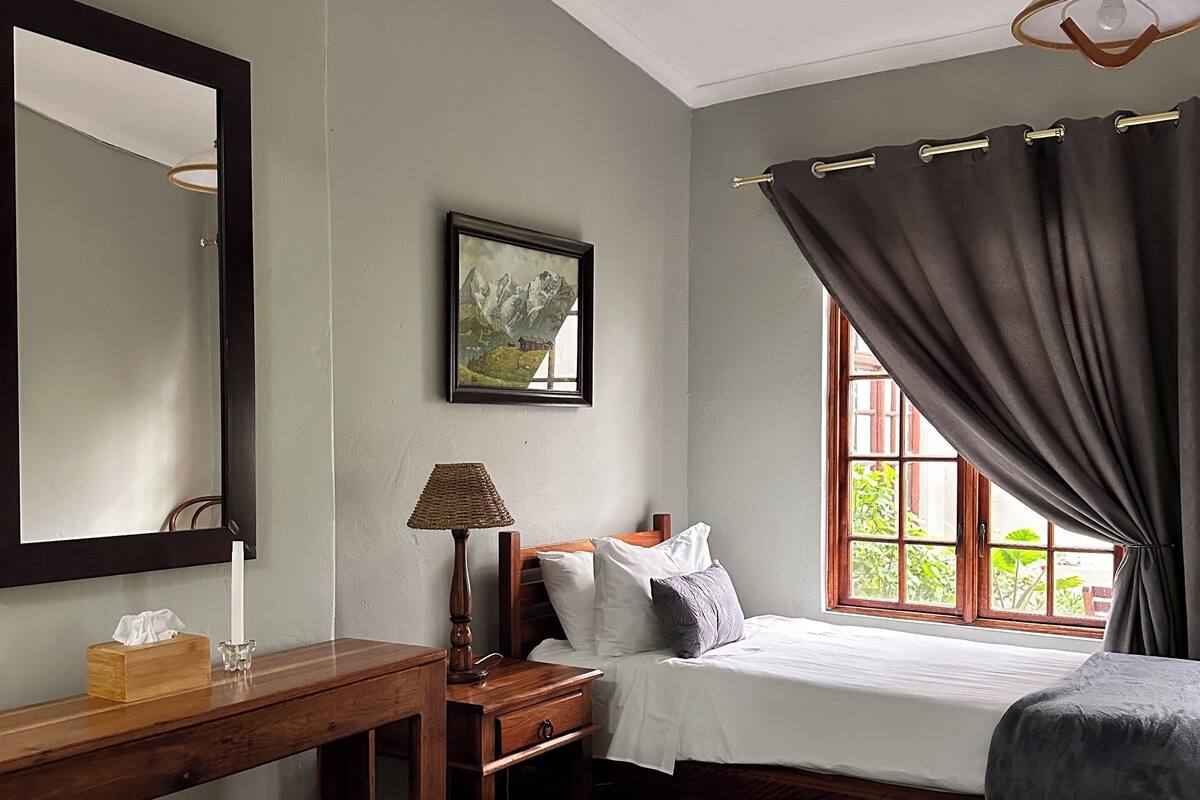 A bedroom with a bed, a bedside table with a lamp, a mirror, and a window with curtains.