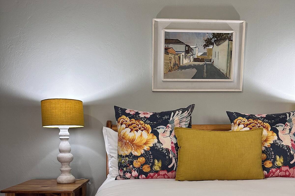 A neatly made bed with a yellow throw blanket, a wooden nightstand with a lamp, and floral-patterned pillows.