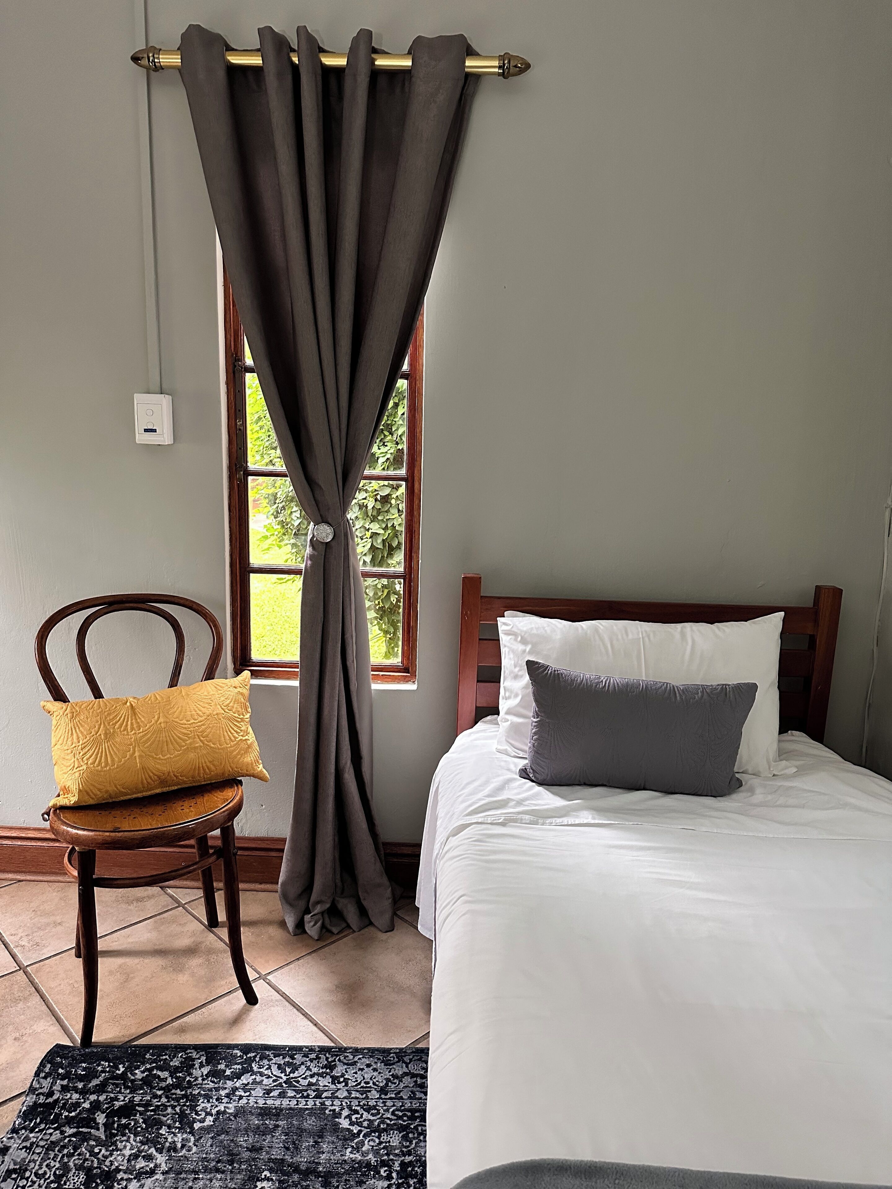 Une chambre à coucher avec un lit, une chaise dotée d’un coussin, une fenêtre avec des rideaux et un thermostat fixé au mur.