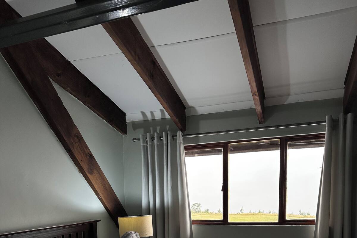 A bedroom with a large bed, wooden beams, a window with curtains, and a nightstand with a lamp.
