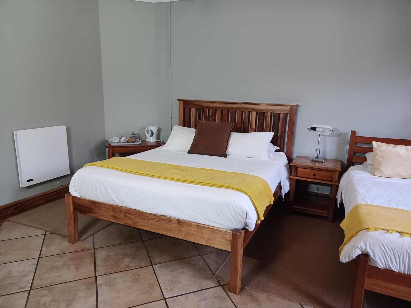 A hotel room with a wooden bed, a bedside table with a lamp, a small table with a kettle, and a radiator on the wall.
