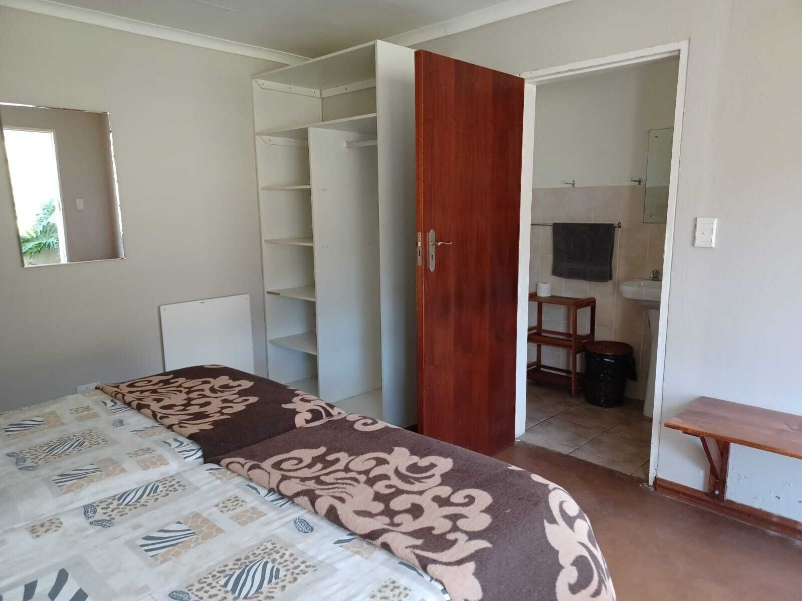 A bedroom with a bed, a wardrobe, a wooden door, a bathroom with a sink and mirror, and a small table.