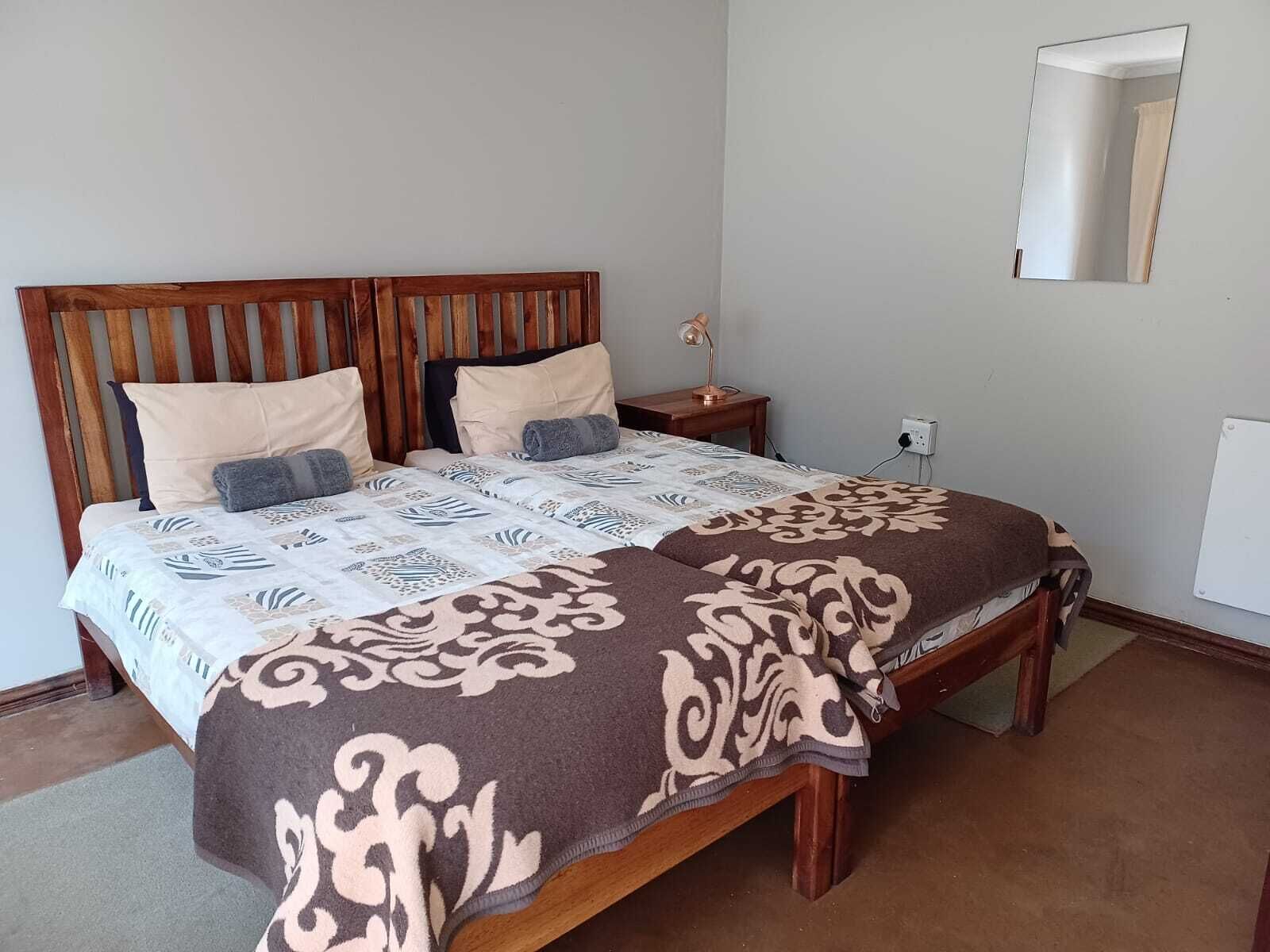 A bedroom with a wooden headboard, two beds with patterned bedding, a bedside table with a lamp, and a wall-mounted mirror.