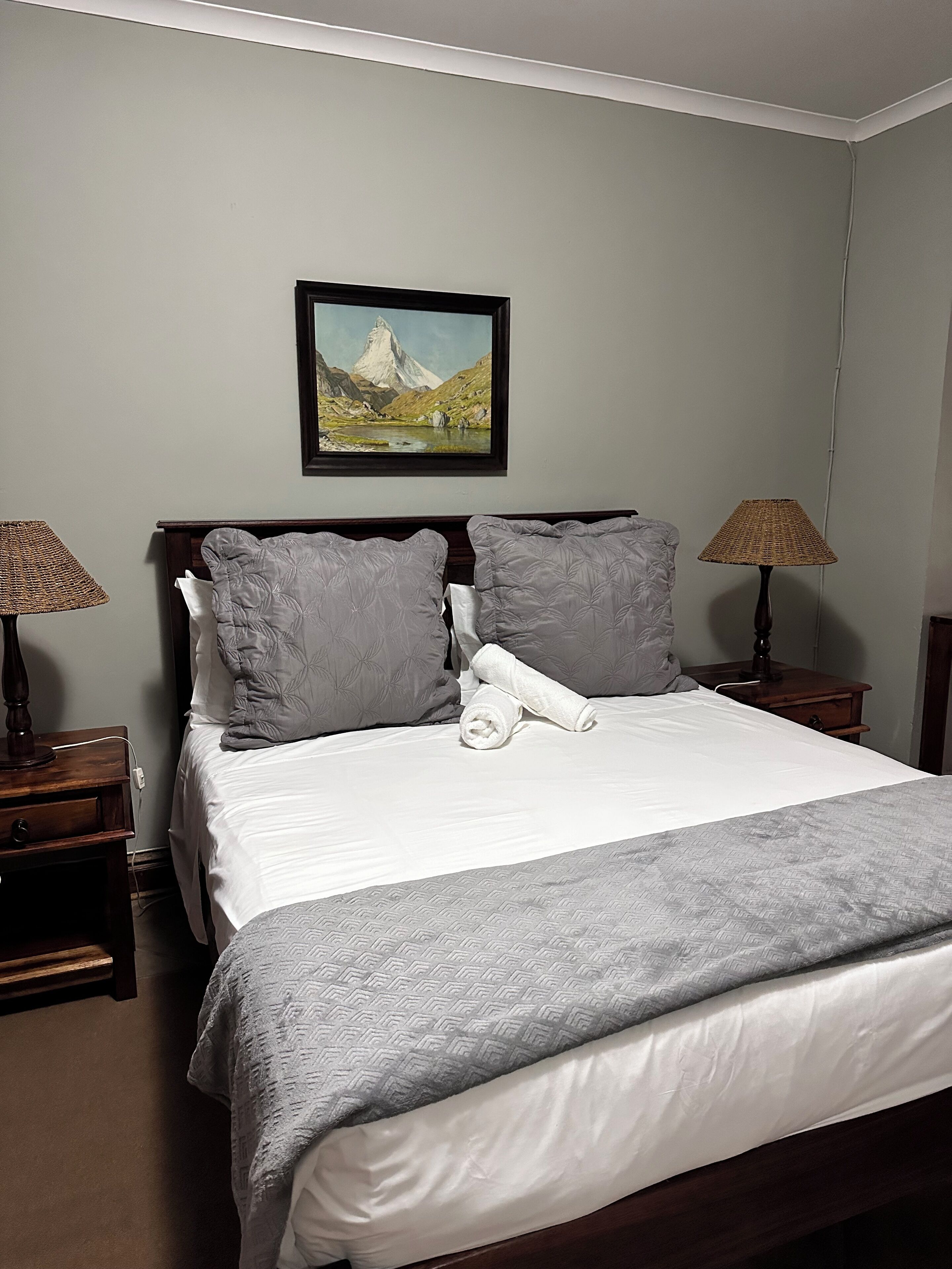 A neatly made bed with a white comforter and gray pillows, a framed landscape painting above the headboard, and two bedside lamps on wooden nightstands.