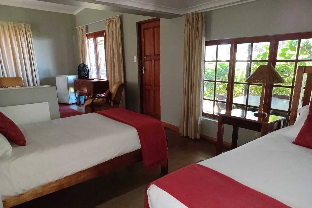A hotel room with two beds, a desk, and a window with a view of greenery.