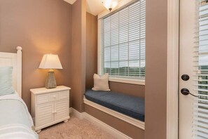 A bedroom with a bed, nightstand, lamp, window seat, and a door with blinds.