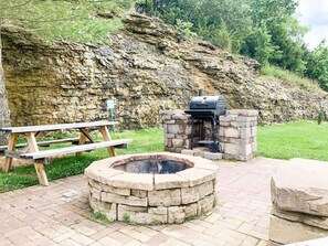 A stone fire pit, a picnic table, and a barbecue grill in a natural setting.