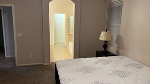 Een slaapkamer met een bed, een nachtkastje met lamp, een raam met zonwering en een boogvormige deuropening naar een andere kamer.