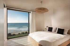 A bedroom with a large window overlooking a beach, a bed with white bedding, and a woven ceiling light fixture.
