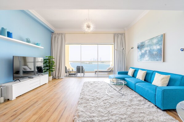 A modern living room with a blue sofa, a TV, a plant, and a view of the sea through large windows.