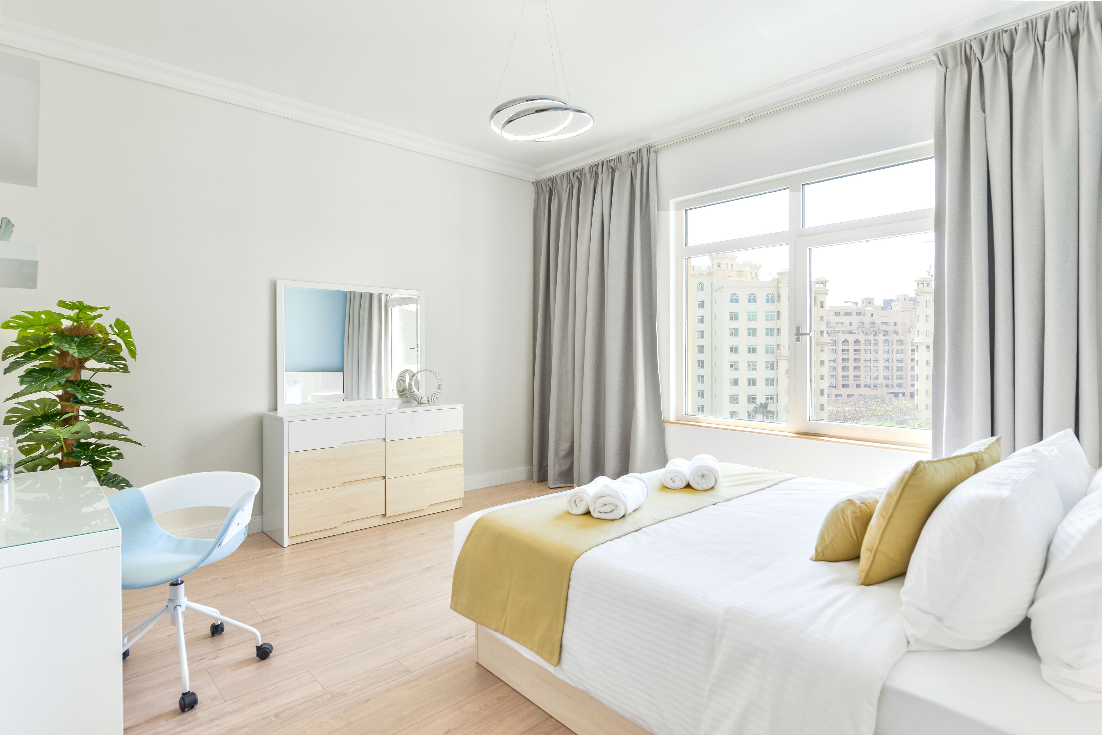 Habitación de hotel moderna con una cama grande, un escritorio con silla, un espejo, una planta y vistas de la ciudad a través de amplios ventanales.