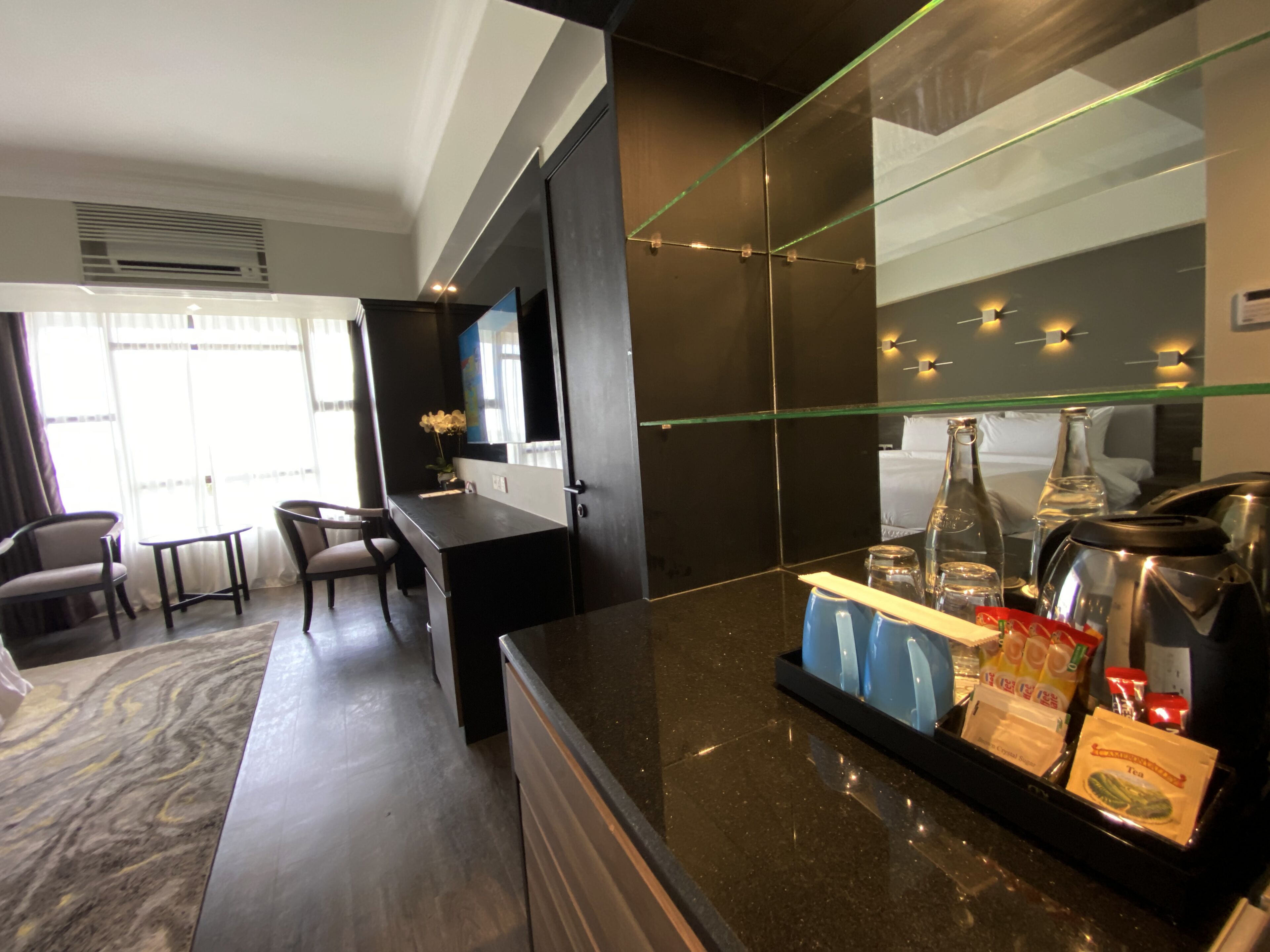 A modern hotel room with a dark-toned kitchen area, a dining table with chairs, and a large window providing natural light.