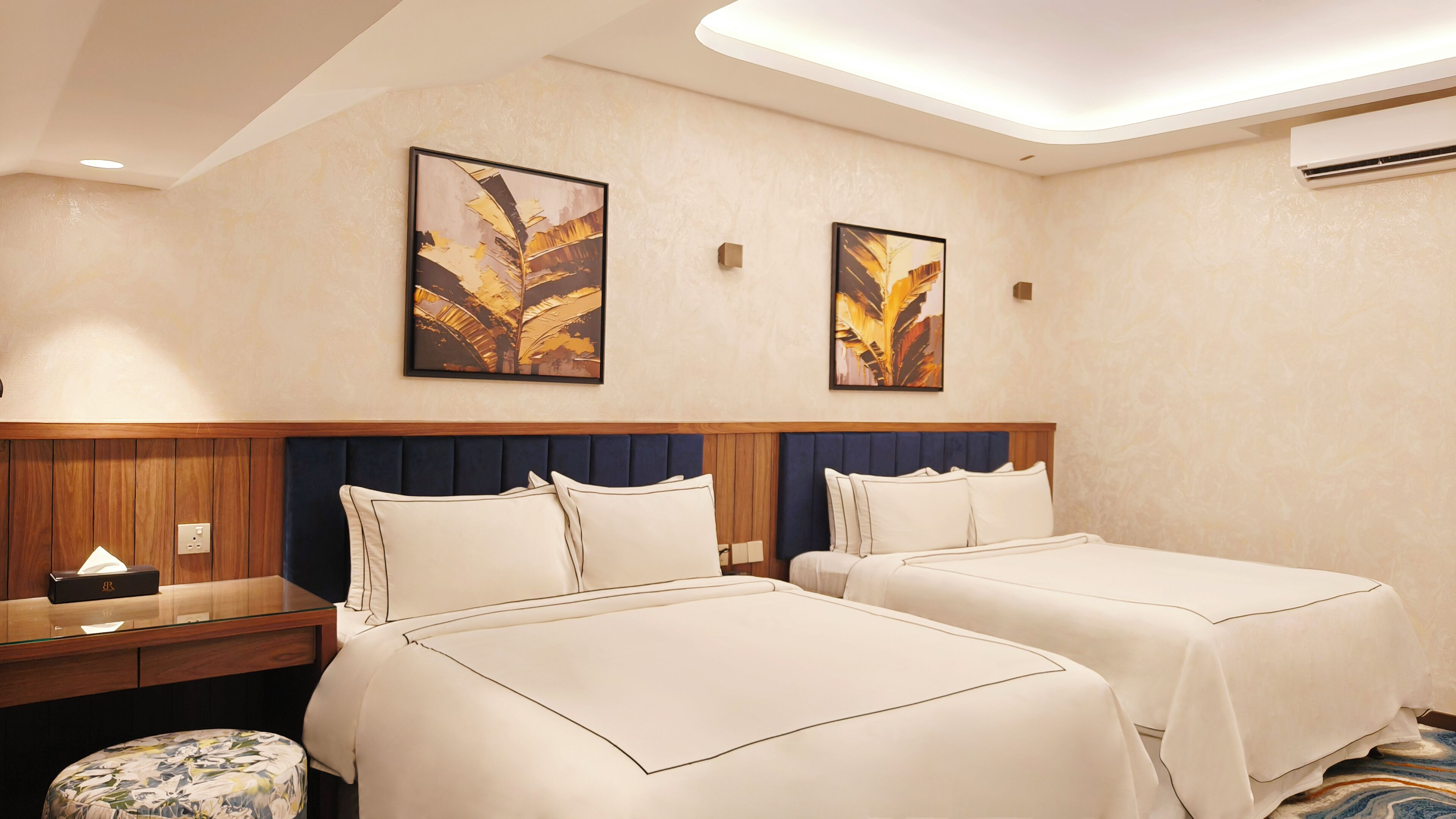Habitación de hotel con dos camas, cabecera de madera, mesita de noche, lámpara y dos cuadros colgados en la pared.