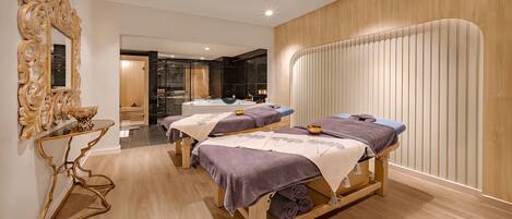 Un cuarto de spa con tres camillas para tratamientos, piso de madera, un espejo grande y un panel de madera en la pared.