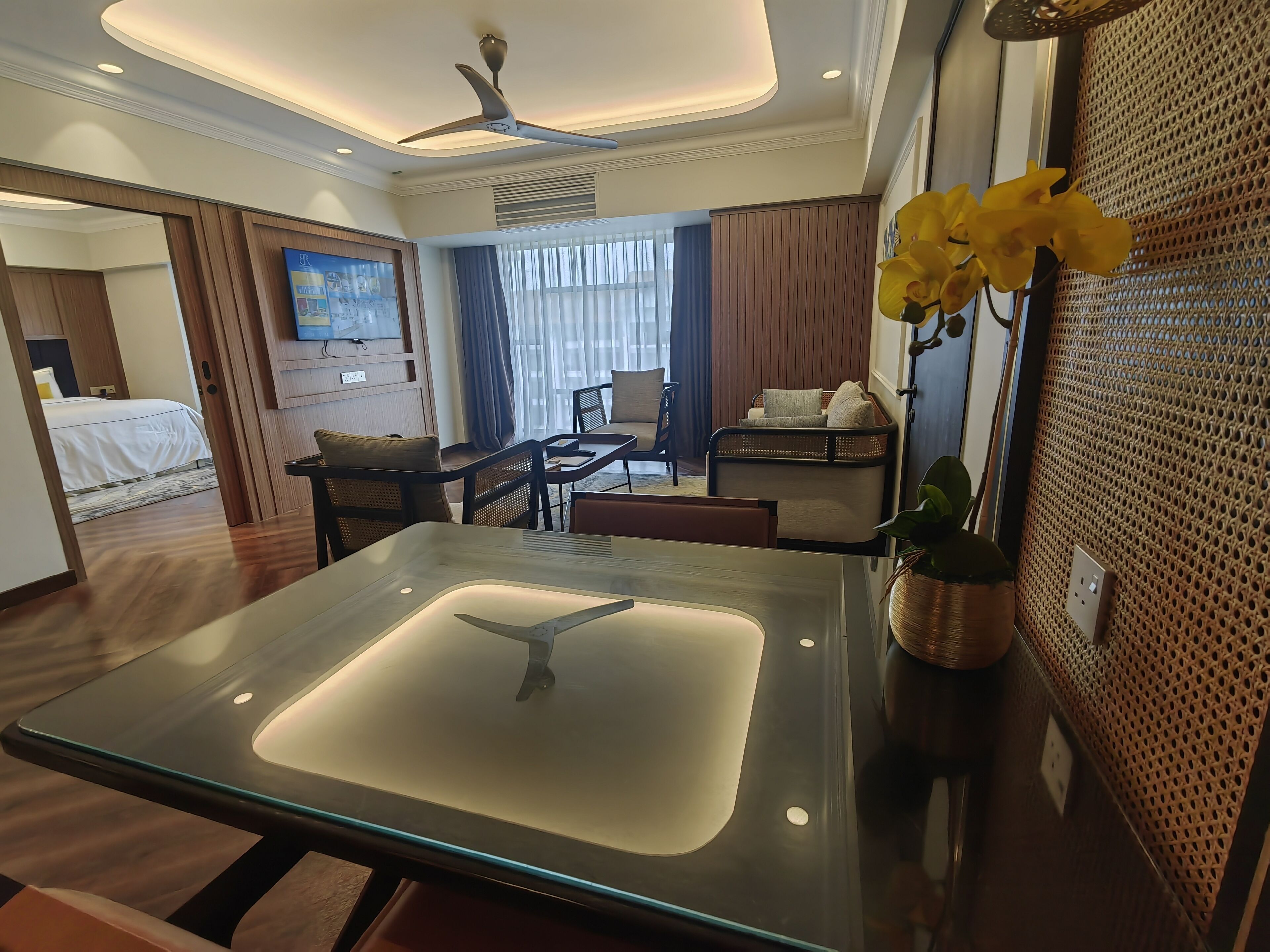 Una habitación de hotel moderna con una mesita de vidrio, ventilador de techo, cama con edredón blanco y zona de estar con sofá y sillones.