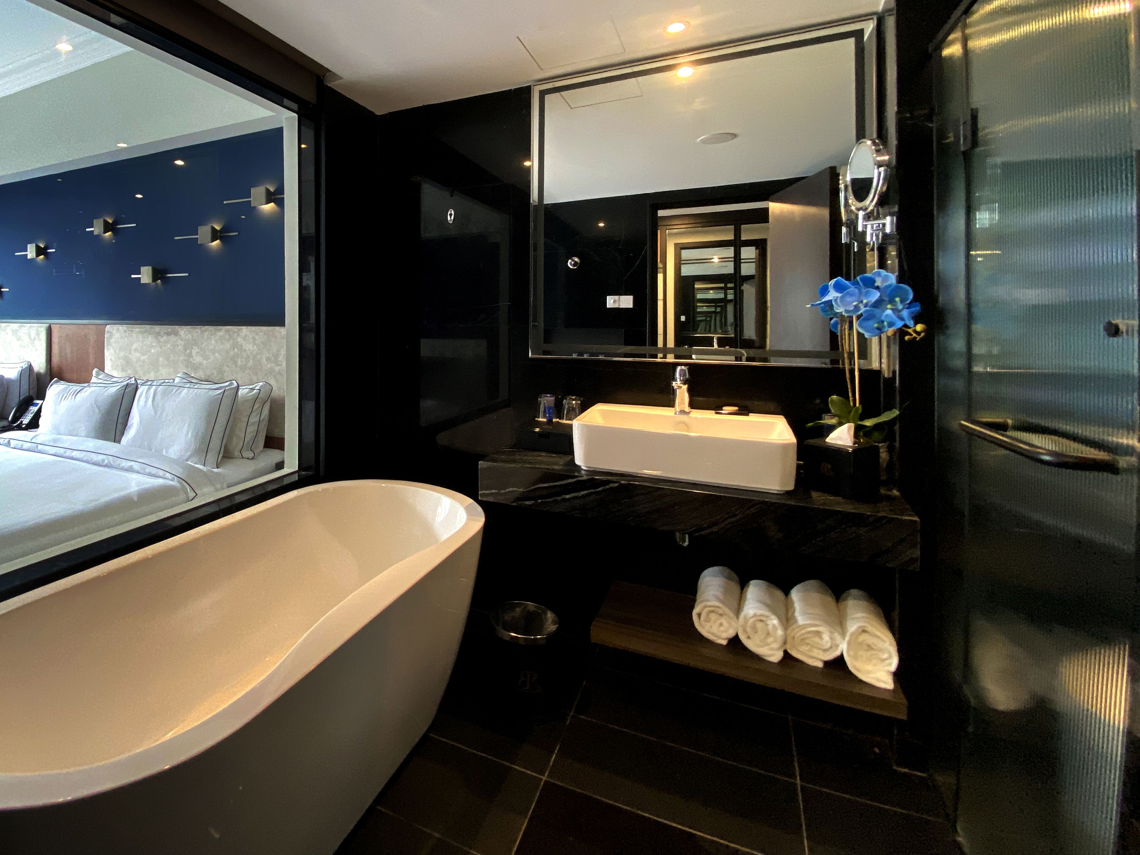 A modern bathroom with a large bathtub, a sink, and a mirror. There are towels on a shelf and a phone on the bed.