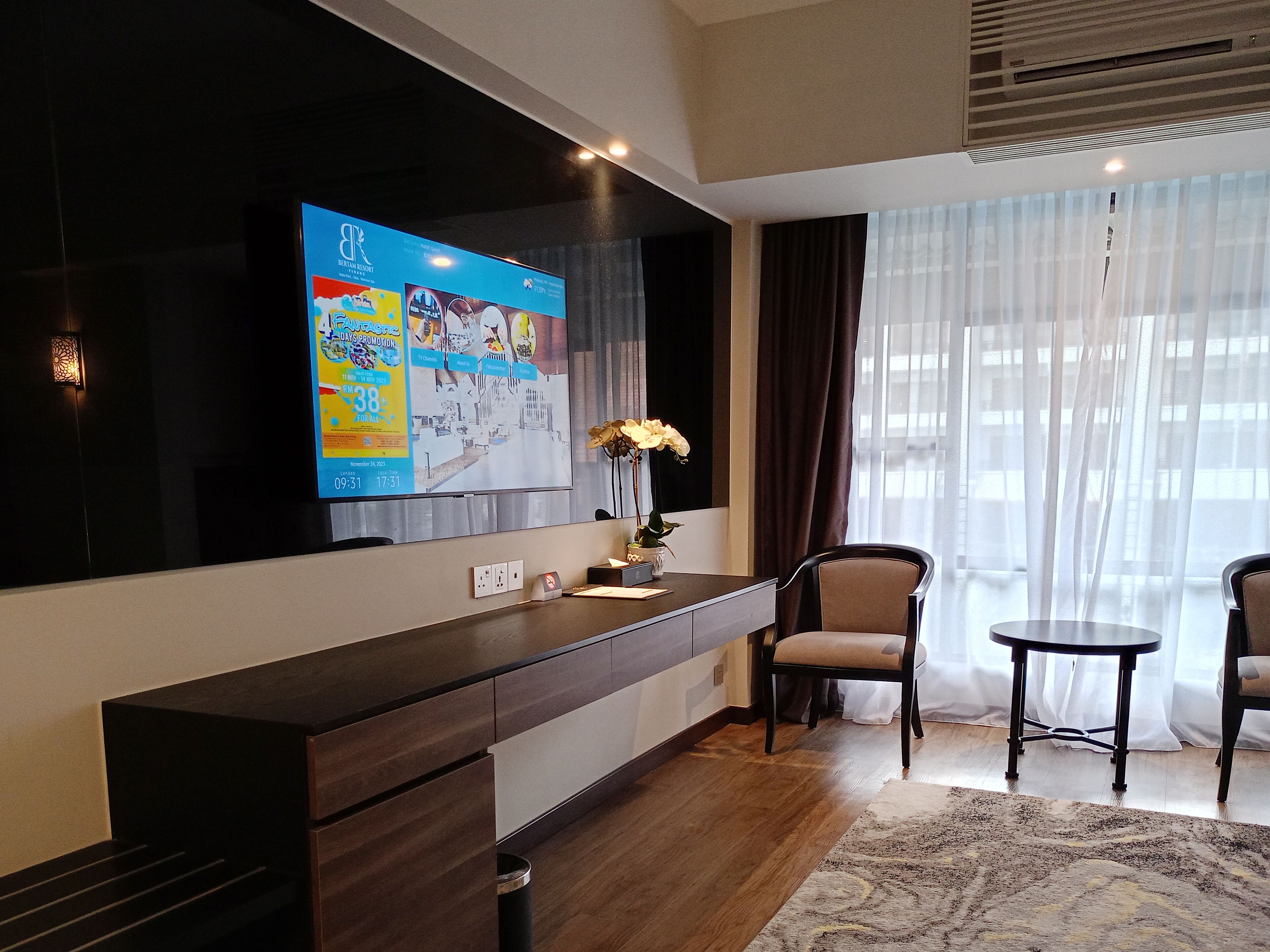 A modern hotel room with a large flat-screen TV mounted on the wall, a wooden console table, two chairs, a small round table, and a large window with sheer curtains.