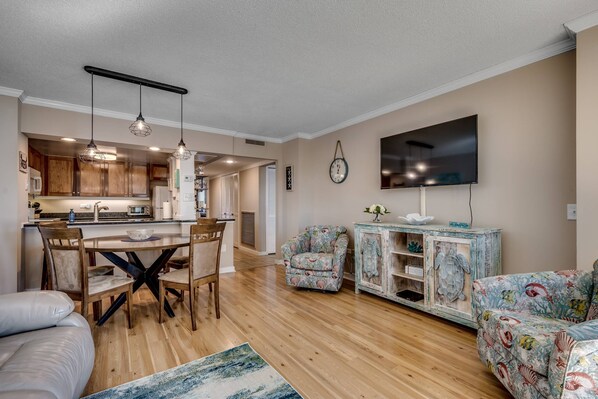 A modern living room with a wooden floor, a dining area with a cross-back table, and a TV mounted on the wall.