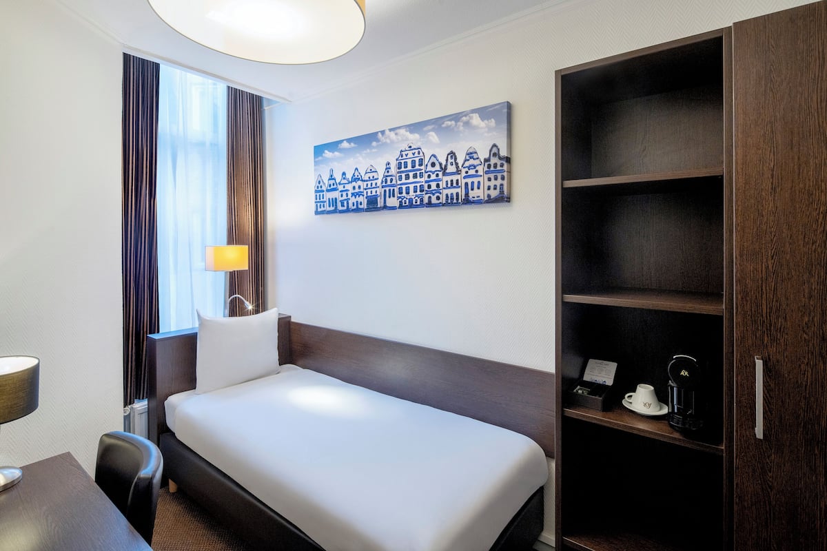 A hotel room with a bed, a desk, a lamp, a coffee machine, and a painting on the wall.