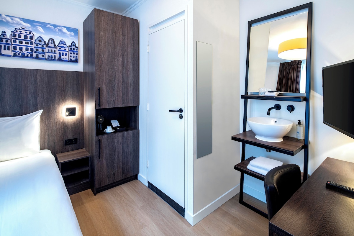 A hotel room with a bed, a bedside table, a built-in wardrobe, a bathroom with a sink, mirror, and towel rack.