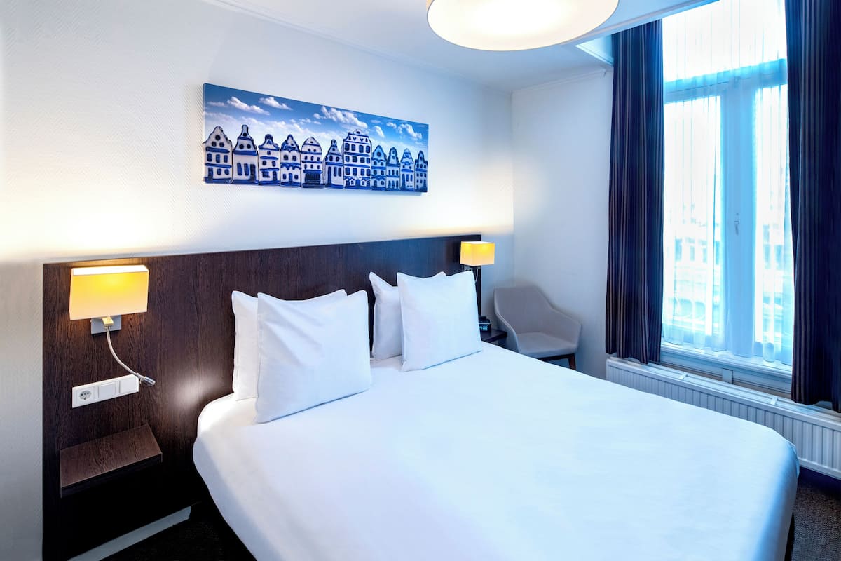 A hotel room with a large bed, two bedside lamps, a nightstand, a chair, a window with curtains, and a wall painting of buildings.