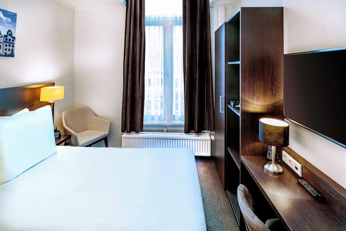 A hotel room with a bed, a chair, a TV, and a window with curtains.