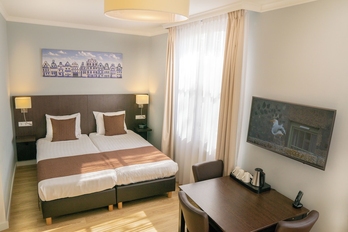 A hotel room with a double bed, bedside tables with lamps, a desk with a chair, a painting on the wall, and a window with curtains.