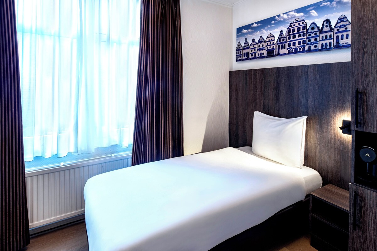 A hotel room with a bed, a window with curtains, a wall-mounted artwork of buildings, and a wooden headboard.