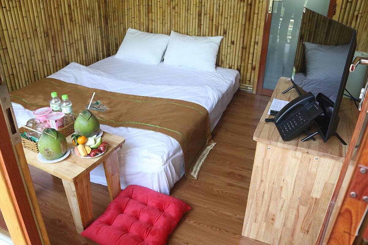 A room with a bed, a desk with a phone, a red cushion, and a table with coconuts and fruits.