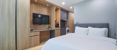 Una habitación de hotel moderna con una cama grande, un televisor de pantalla plana montado en la pared, una cocina integrada con microondas y lavadora, y paneles de madera en las paredes.