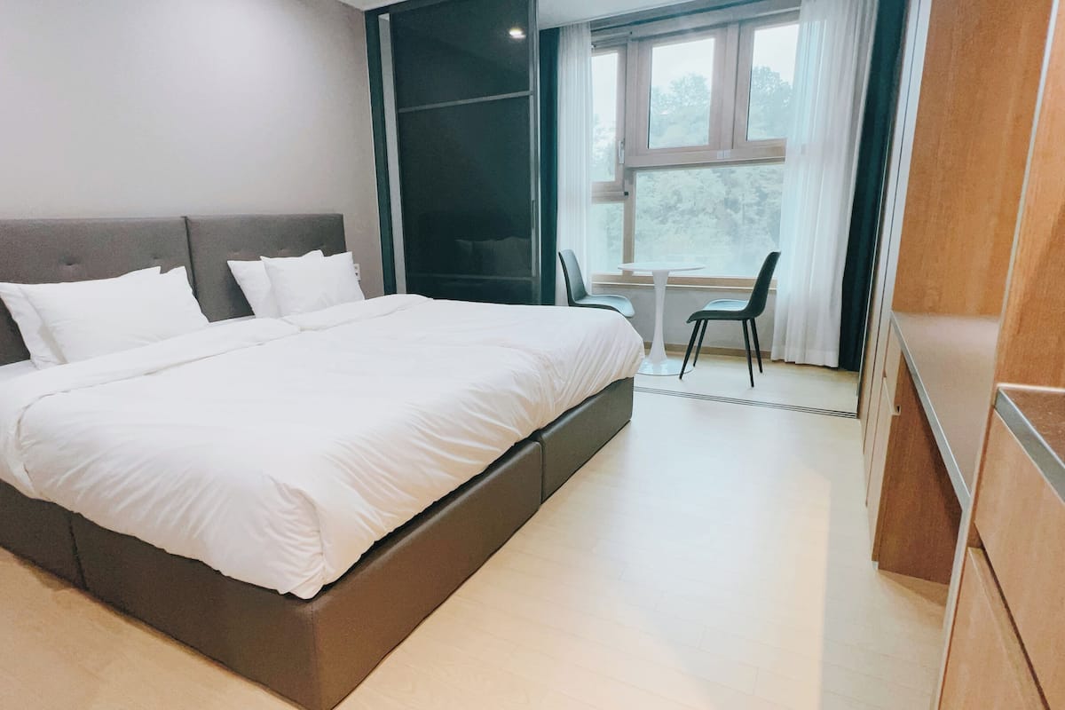 Ein modernes Schlafzimmer mit einem großen Bett, einem kleinen Tisch, zwei Stühlen und Blick ins Freie durch ein Fenster.