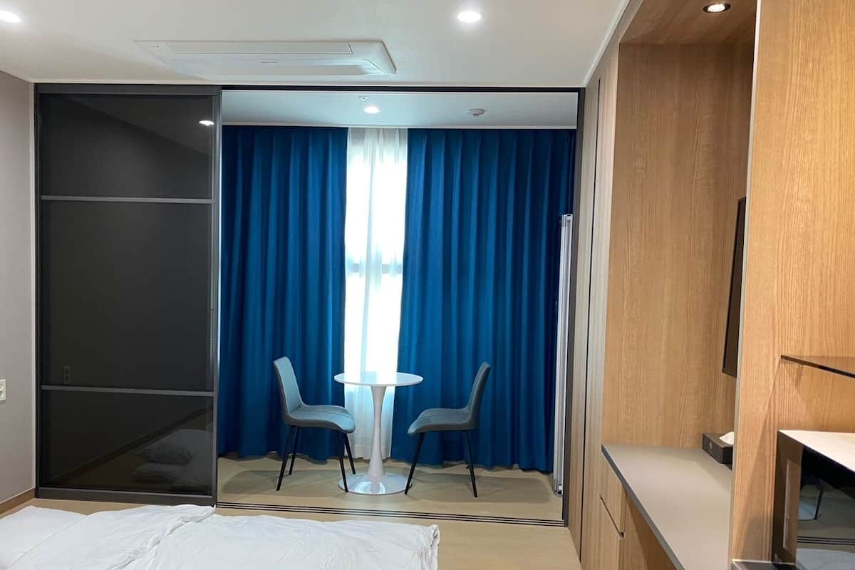 Ein Hotelzimmer mit einem Bett, einem Schreibtisch, zwei Stühlen und einem Fenster mit blauen Vorhängen sowie einer Schiebetür.