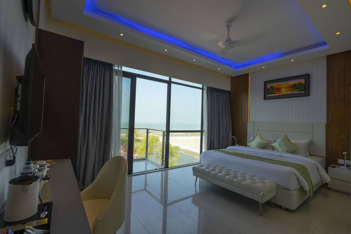 A hotel room with a large bed, a desk with a chair, a television, and a balcony with a view of the beach.