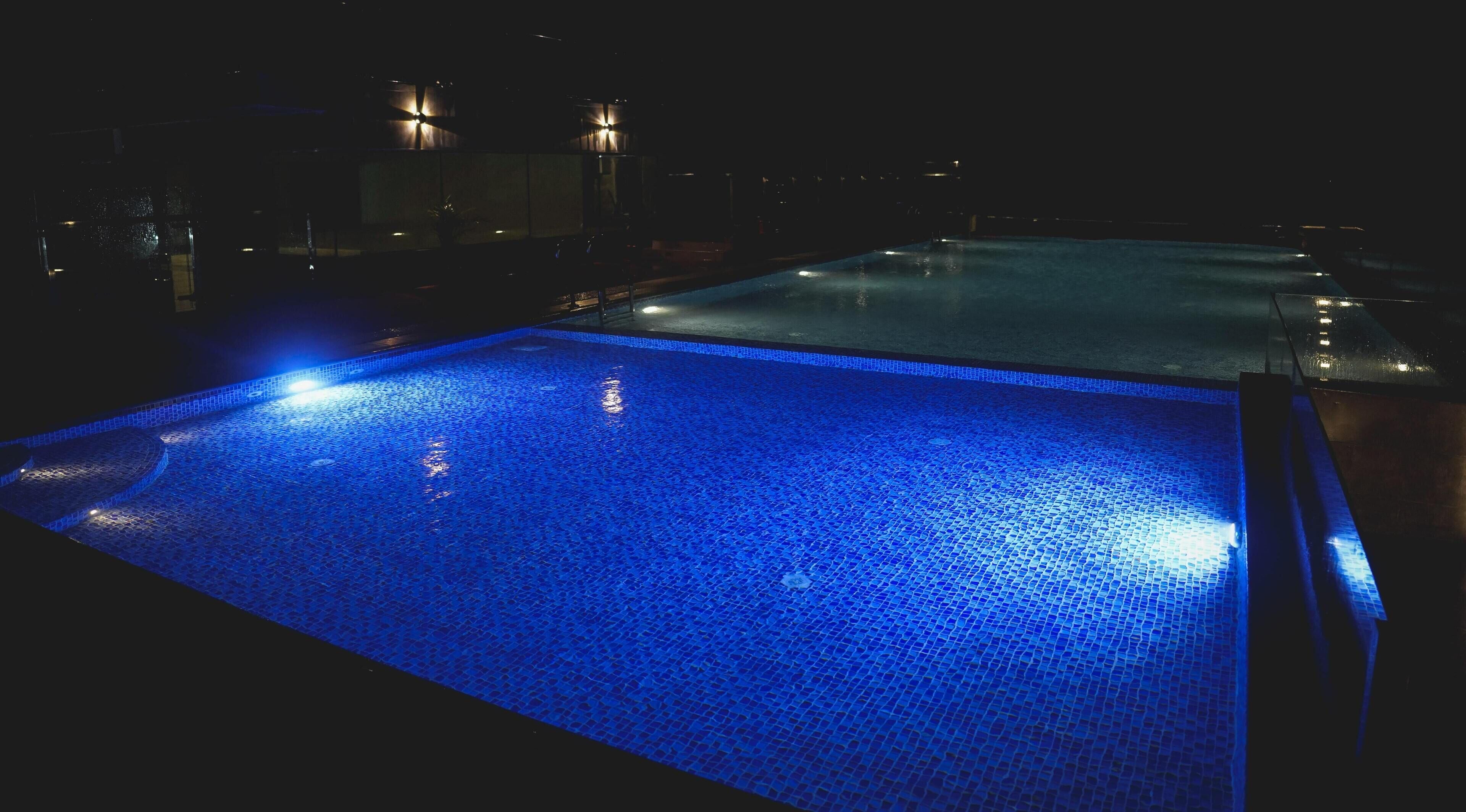 A nighttime view of a swimming pool with blue lighting, surrounded by dark areas and some distant lights.