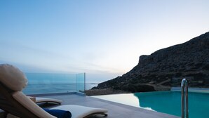 A poolside area with lounge chairs, a glass barrier, and a view of the sea and rocky terrain. - Krystallon West - Superb sea views, walking distance to the beach and Paleochora (Chania)