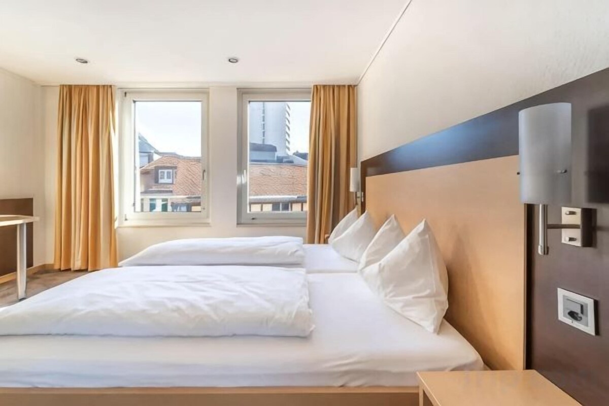A hotel room with a large bed, a bedside table, a lamp, and a window with curtains.