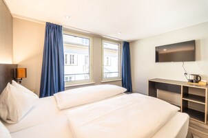 Habitación de hotel con cama, escritorio, televisión y ventanas amplias con cortinas.