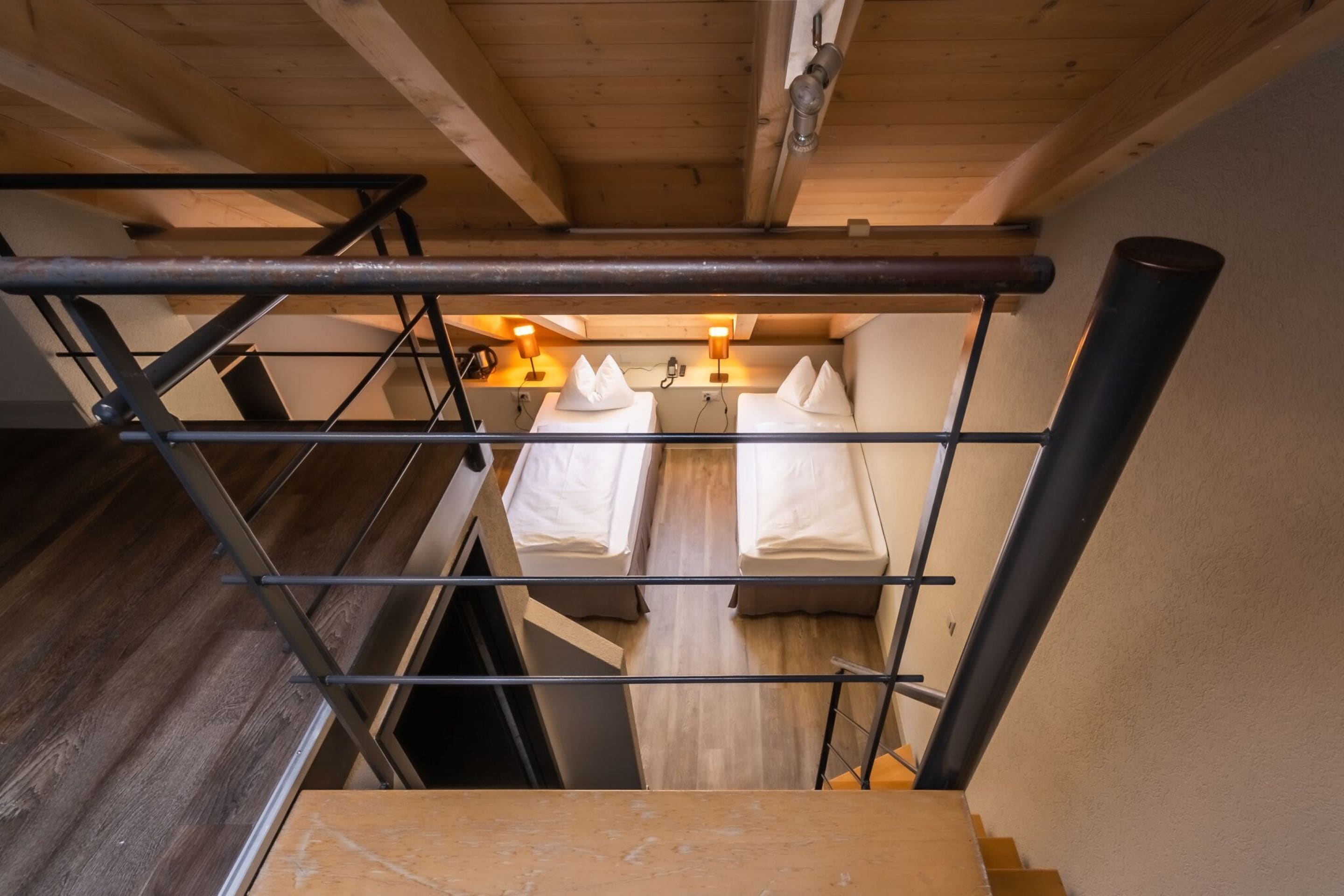 Un escalier mène à une pièce où se trouvent deux lits, chacun étant équipé d’un oreiller et d’une lampe fixée au mur.