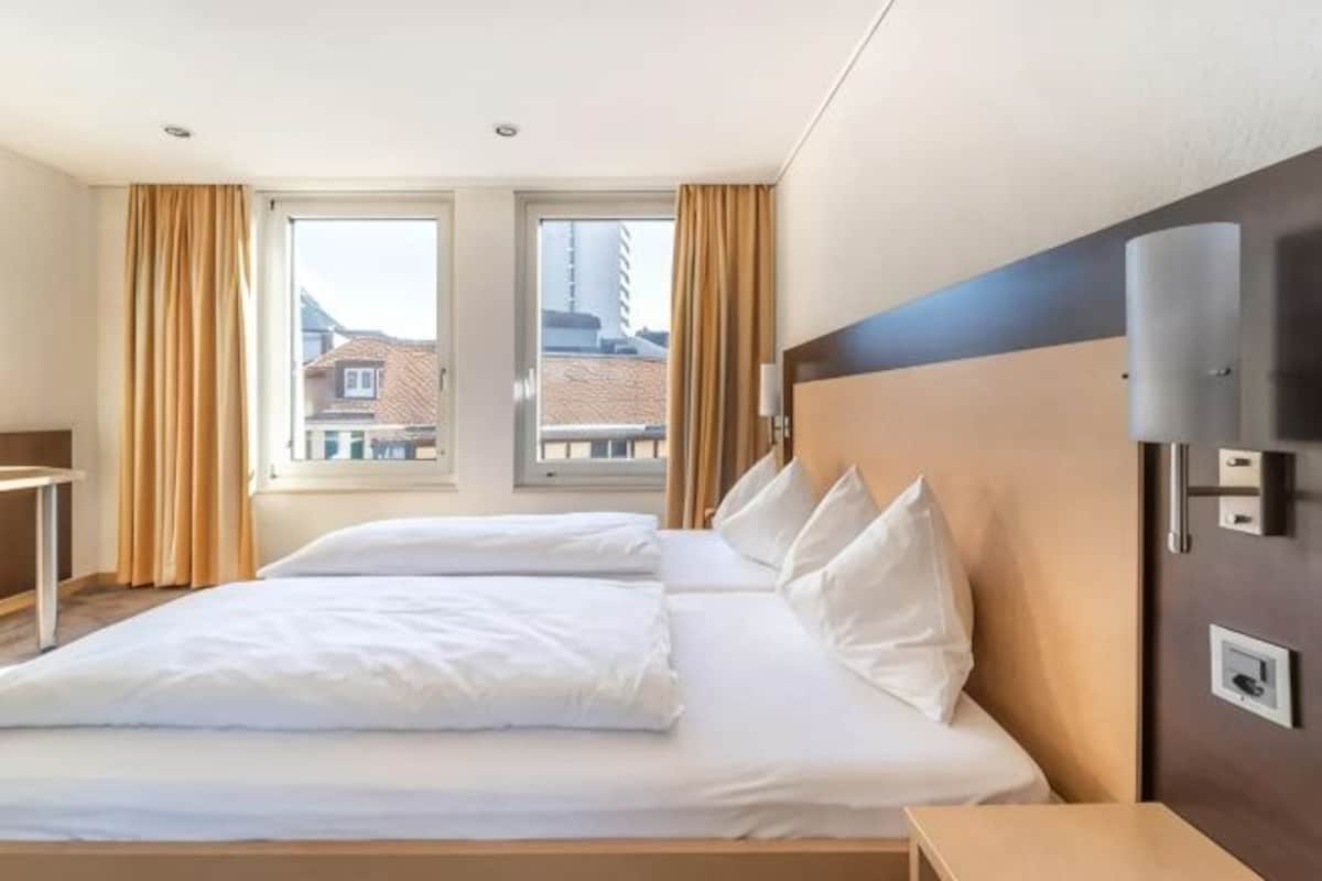 A hotel room with a large bed, a bedside table, a lamp, and a window with curtains.