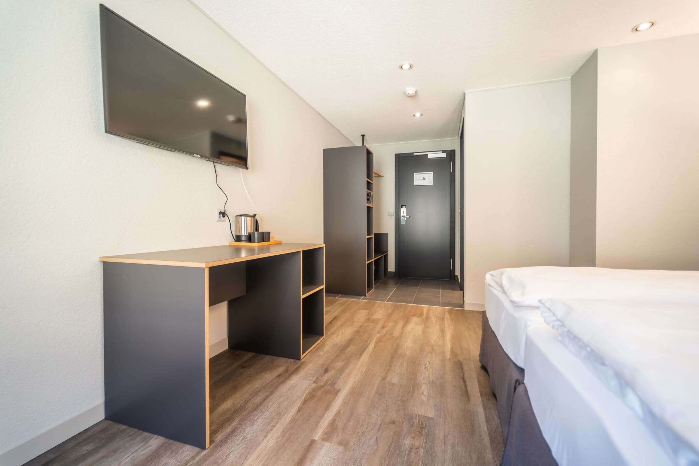 A modern hotel room with a wooden desk, a bed, a flat-screen TV, and built-in shelving.