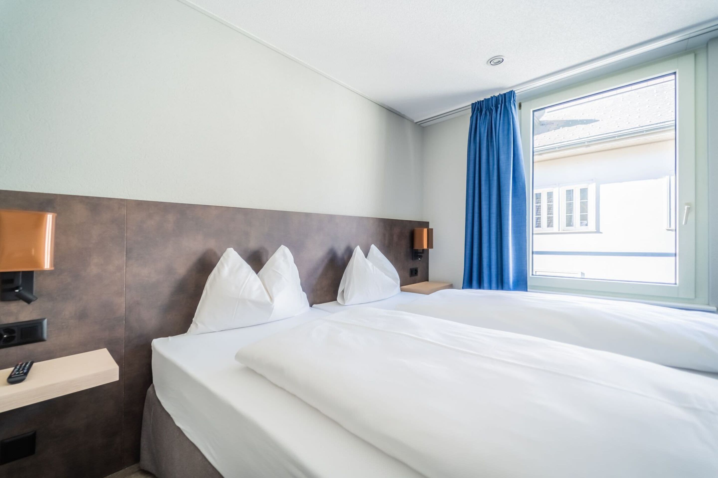 Une chambre d’hôtel avec un grand lit, une table de chevet avec une lampe, une petite table de nuit avec un téléphone et une fenêtre avec des rideaux bleus.
