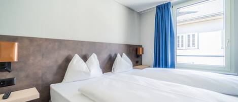 A hotel room with a large bed, a bedside table with a lamp, a nightstand with a phone, and a window with blue curtains.